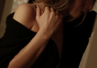 Woman in a black off-the-shoulder sweater holding the back of her neck, face turned away, in dim warm light.